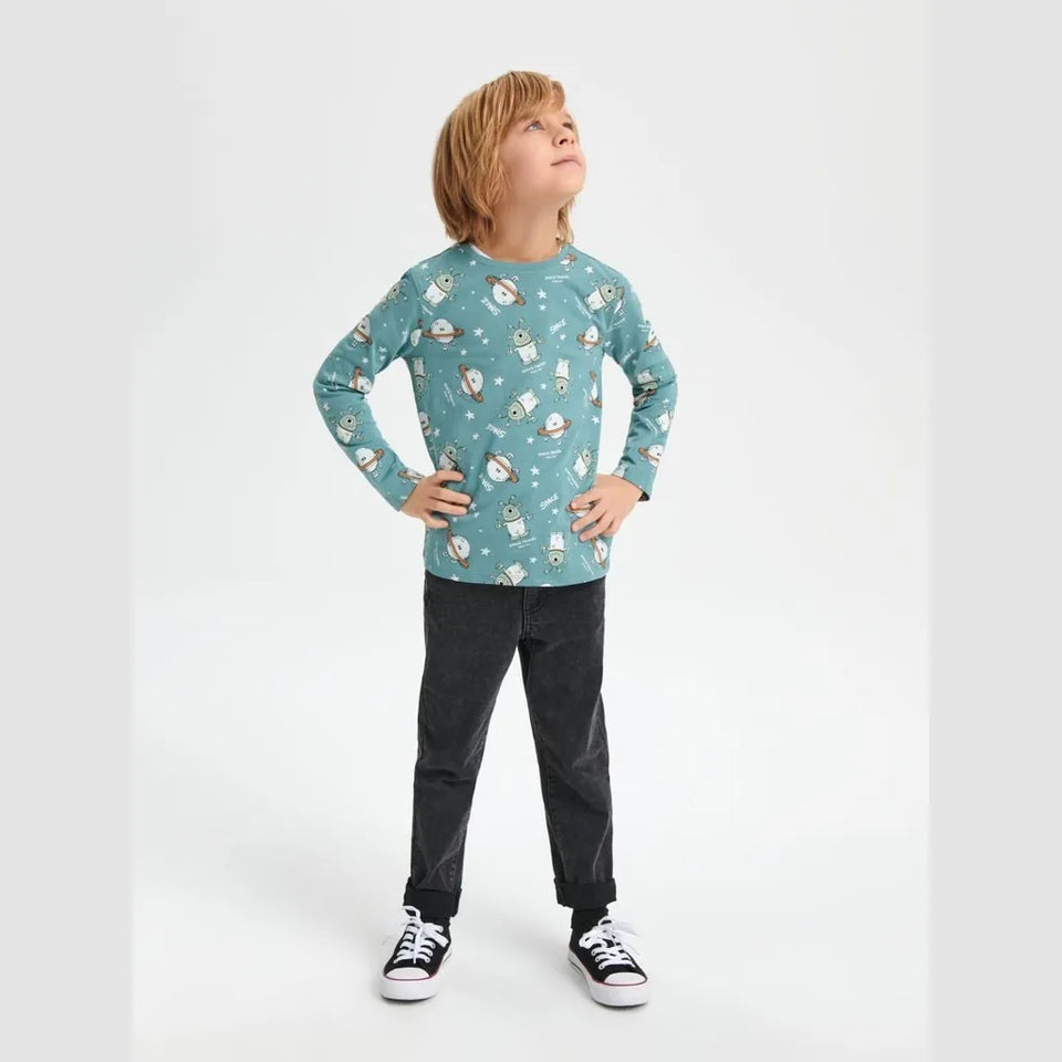 A young boy with blonde hair stands in a studio wearing a teal long-sleeve t-shirt with a snowman pattern, dark gray jeans, and black sneakers with white soles, posing with hands on hips against a light gray background.