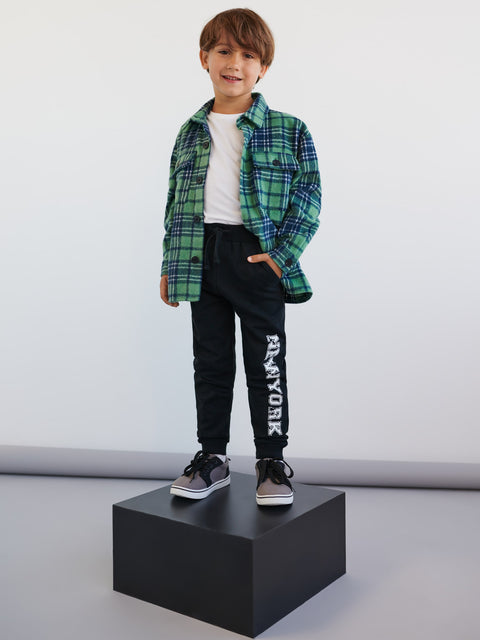 Child wearing a green plaid shirt, black pants with 'New York' text, and black shoes on a white background