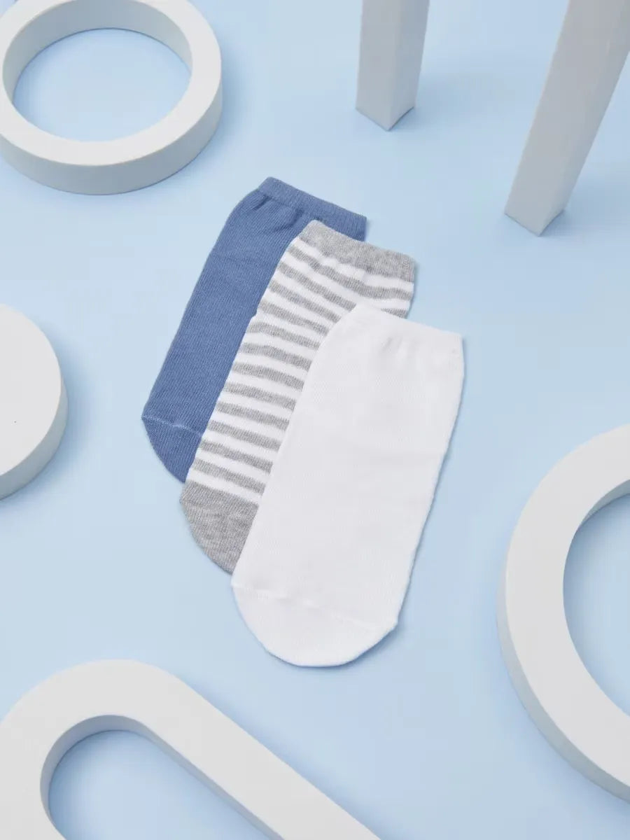 Three pairs of low-cut ankle socks arranged in a fan shape on a light blue background with white geometric props. The socks are white, heather gray with thin gray stripes, and medium blue.