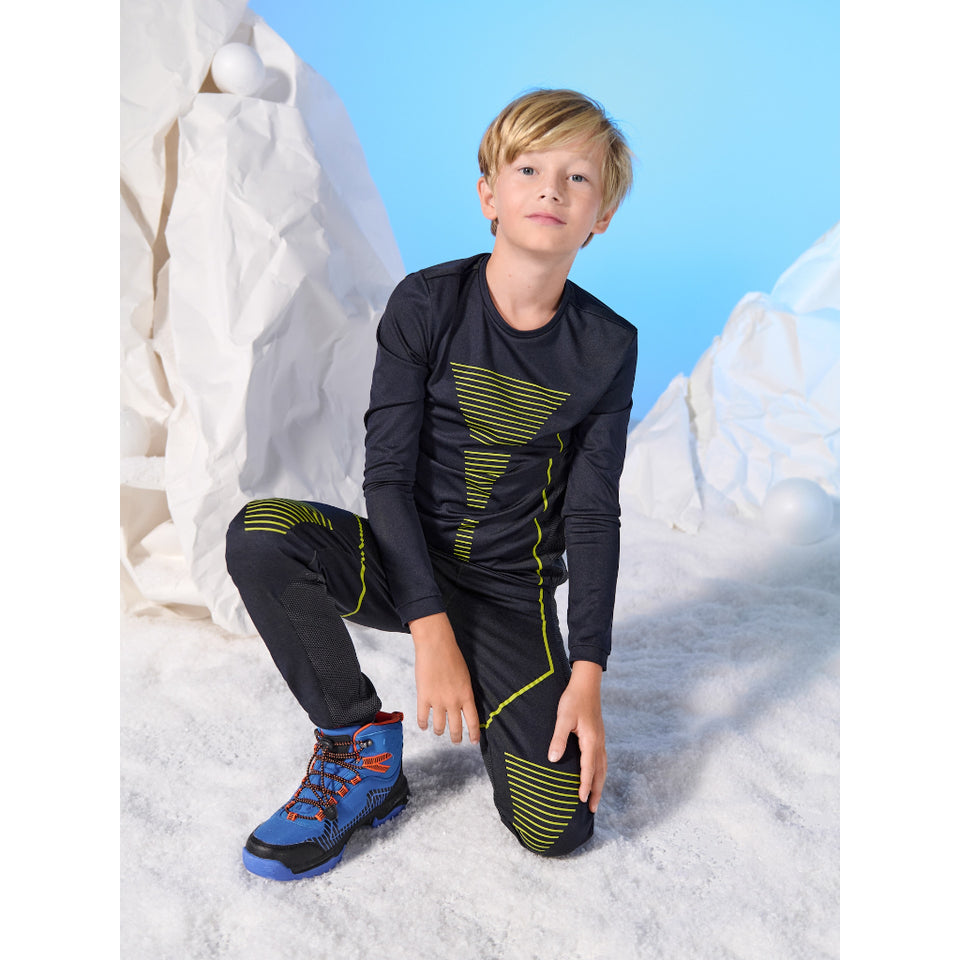 A young boy wearing a matching black athletic long-sleeve shirt and pants set with lime green accents, posing in a snowy, icy outdoor setting with a blue sky background.