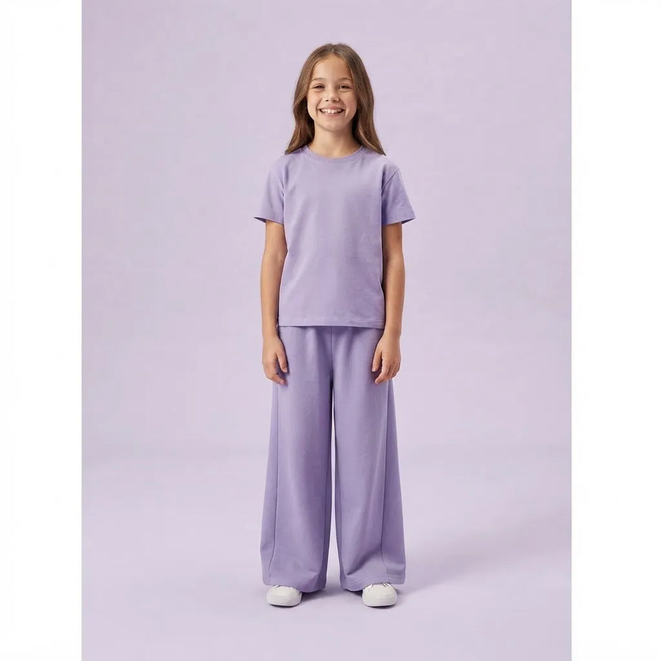 A young girl stands against a solid light purple studio background wearing a matching two-piece lounge set consisting of a short-sleeve crewneck t-shirt and wide-leg pants in a soft lavender color.