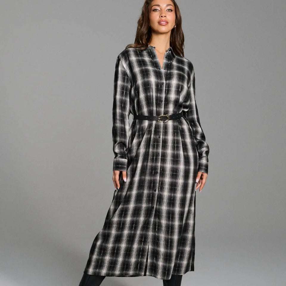 A woman modeling a long-sleeve black and white plaid midi shirt dress with a matching self-tie belt, photographed against a solid gray studio background.