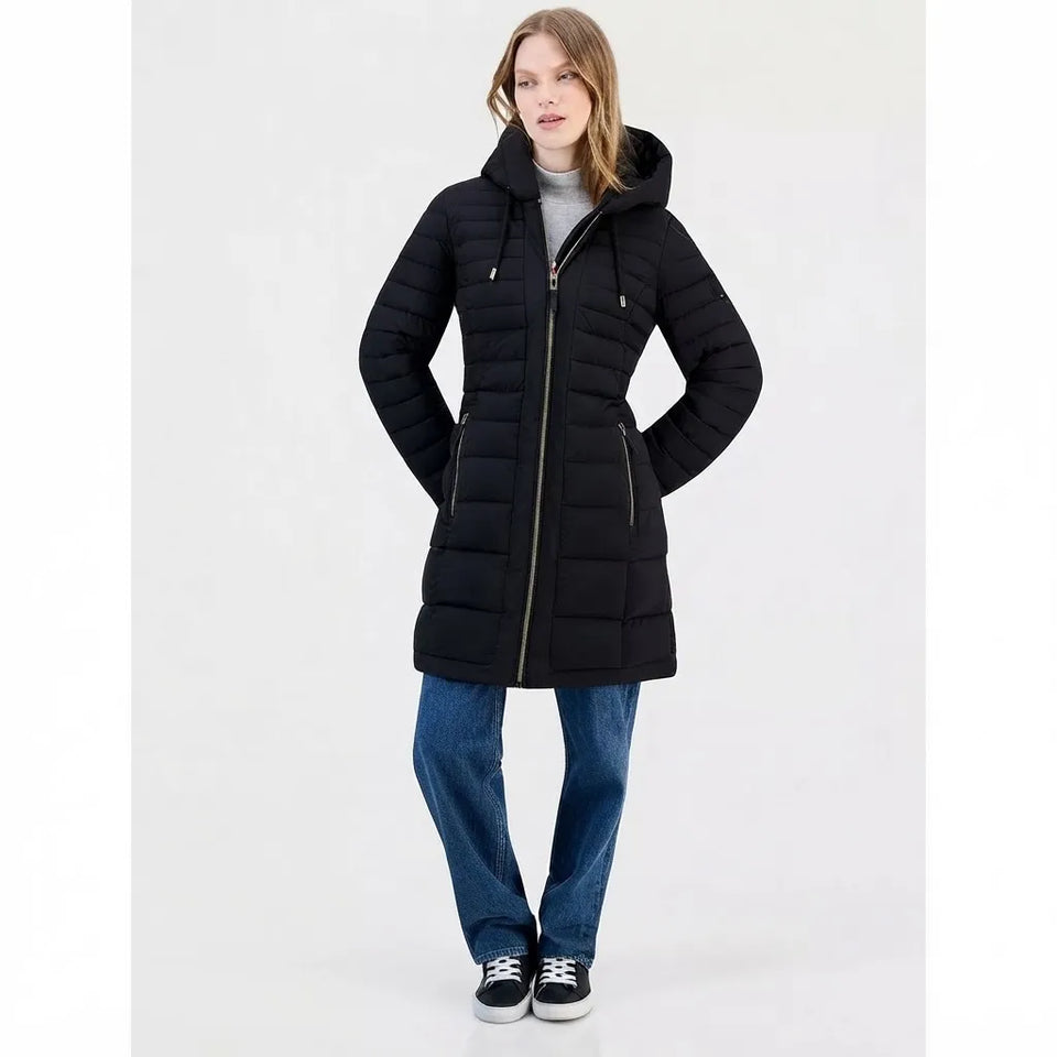 A woman modeling a black hooded puffer coat with gold-toned zippers, paired with blue jeans and black sneakers, against a plain light gray studio background.