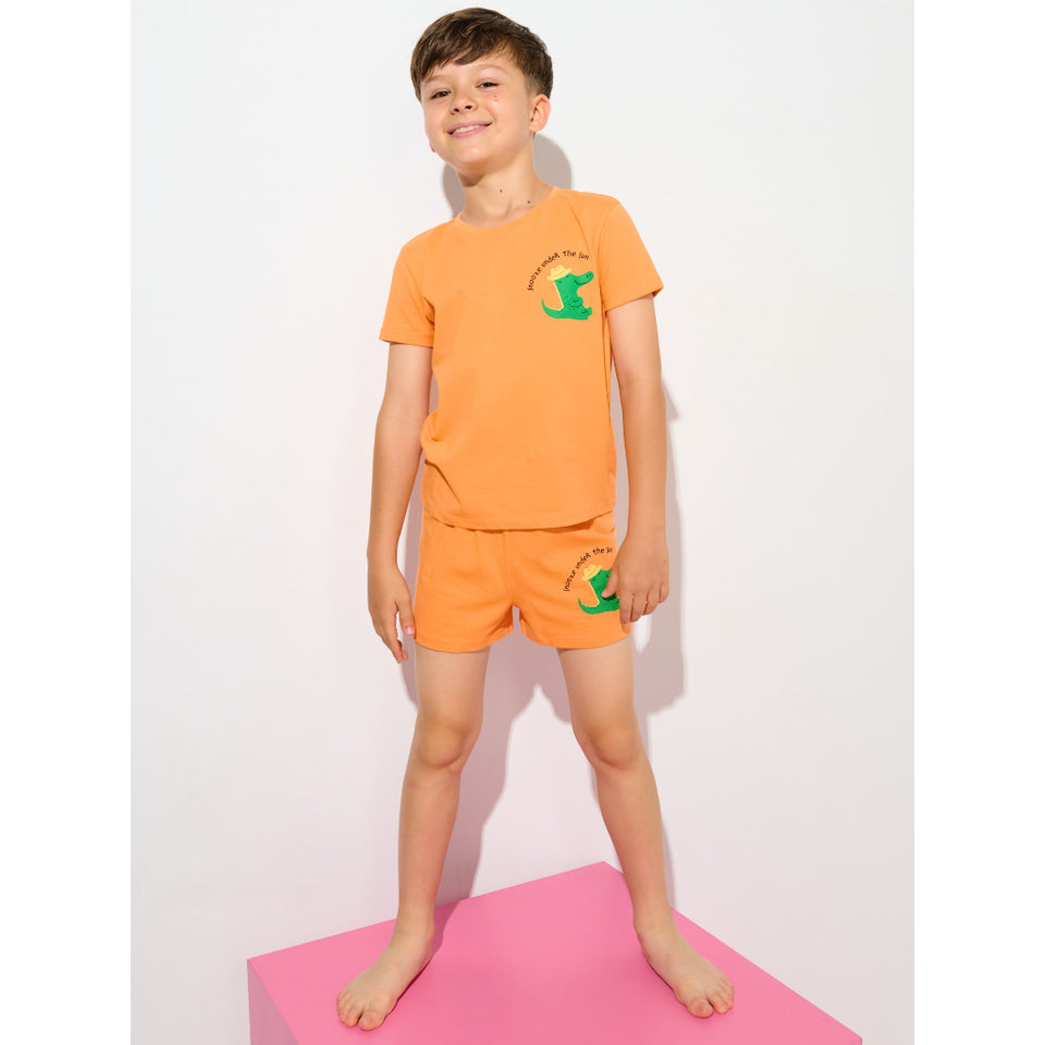 A young boy stands barefoot on a pink block wearing a bright orange two-piece pajama set with a green kangaroo graphic and text on the chest and shorts, against a plain white studio background.
