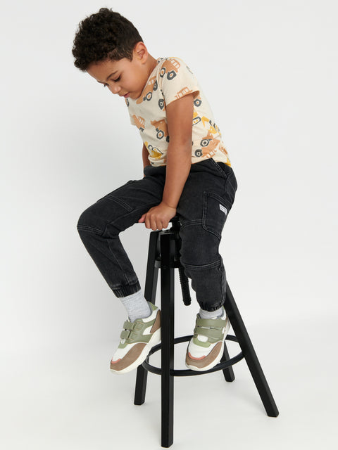 Child sitting on a black stool wearing a patterned shirt and dark pants against a white background