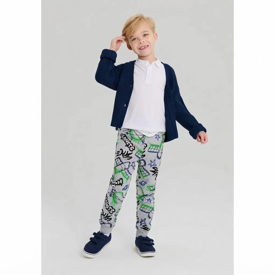 A young boy modeling a casual outfit consisting of a navy blue cardigan, a white polo shirt, light gray pants with a green and black graphic print, and navy blue sneakers, set against a plain light gray studio background.