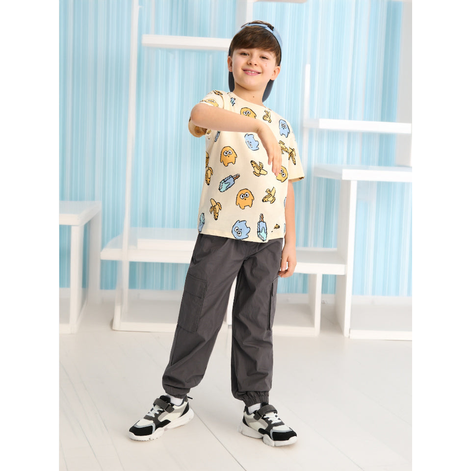 A child model wearing a cream-colored t-shirt with a colorful cartoon character print, dark gray jogger pants, and black-and-white sneakers, posing in front of a light blue and white shelving background.