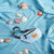 A light blue fanny pack with black straps and a tan leather patch, styled on a draped light blue fabric background with scattered seashells.