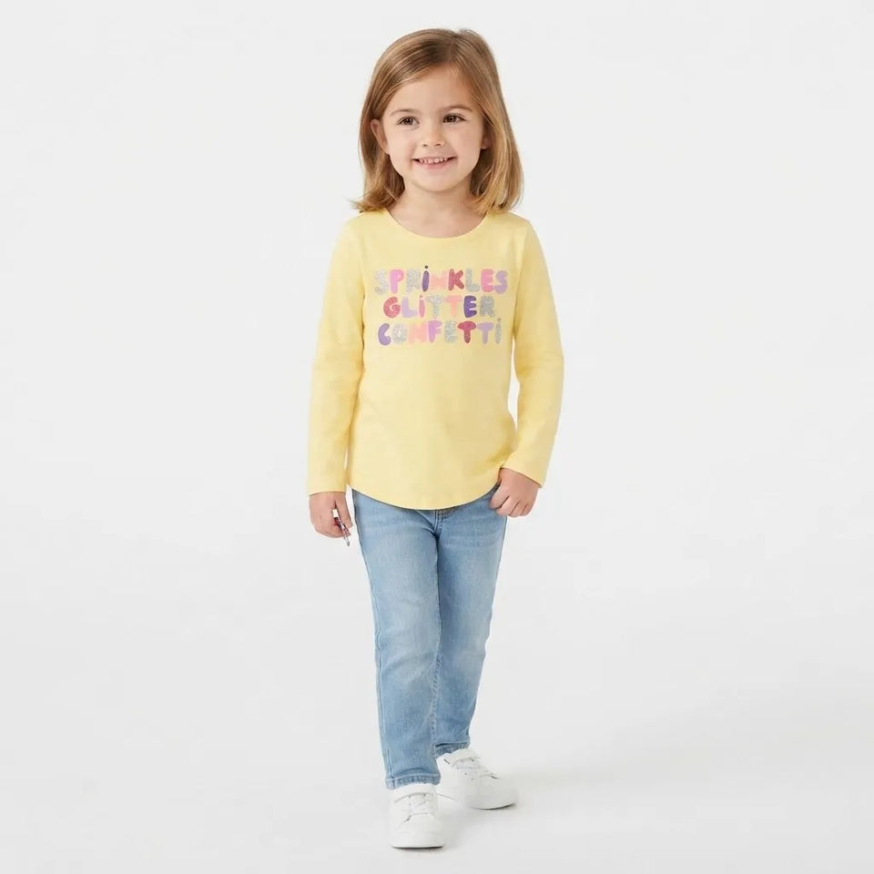 A young girl with light brown hair stands against a white studio background wearing a pale yellow long-sleeve t-shirt with 'SPRINKLES GLITTER CONFETTI!' printed in multicolored letters, paired with light blue jeans and white sneakers.