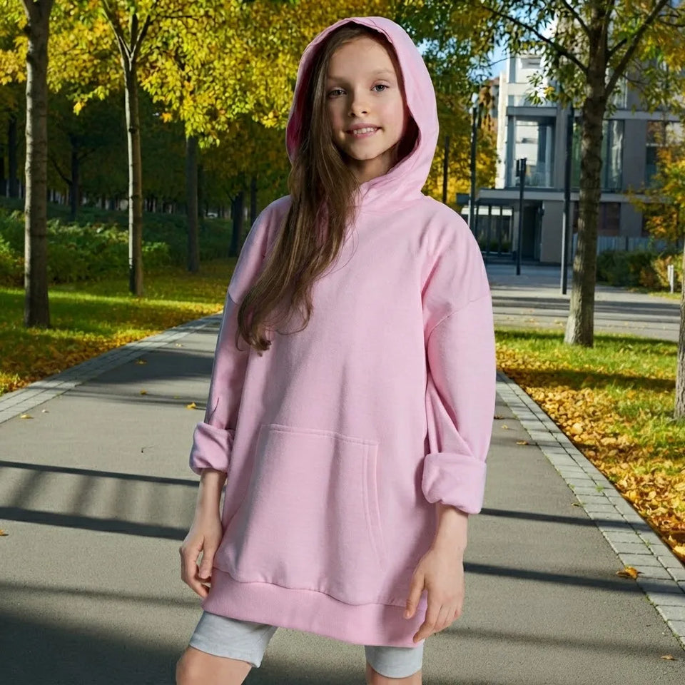 A young girl wearing a light pink oversized hooded sweatshirt with rolled-up sleeves and a front kangaroo pocket, standing on a paved path in an outdoor park setting with autumn trees.