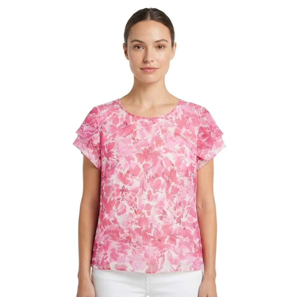 A woman modeling a short-sleeve top with a pink and white abstract floral pattern, paired with white pants, against a plain white background.