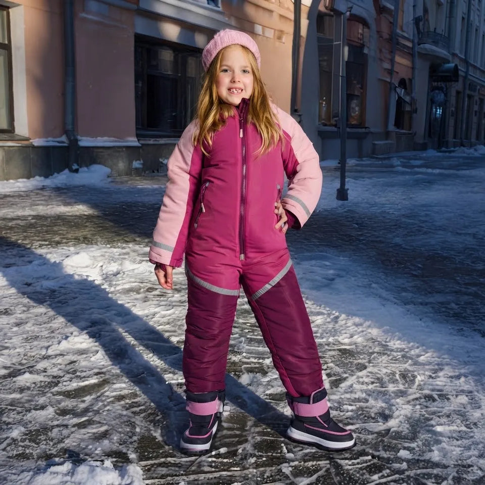 A young girl stands outdoors on a snowy city street wearing a pink and magenta winter snowsuit with a matching pink beanie and dark snow boots.