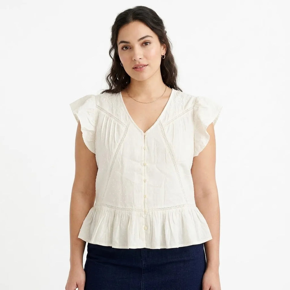 A woman wearing a white sleeveless blouse with ruffled cap sleeves, a V-neckline, and a peplum hem, paired with dark blue jeans against a plain white background.