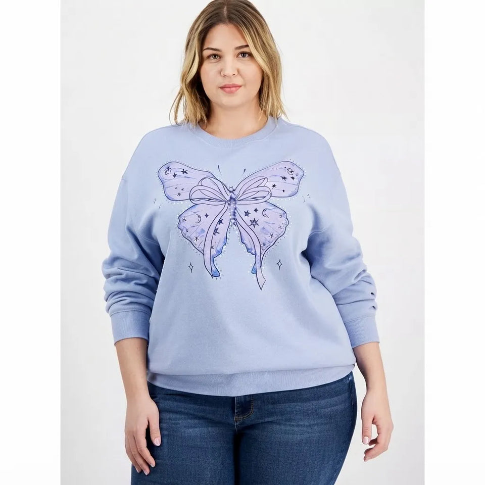 A woman modeling a light blue crewneck sweatshirt with a large lavender and purple butterfly graphic on the front, paired with dark blue jeans, against a plain light gray background.