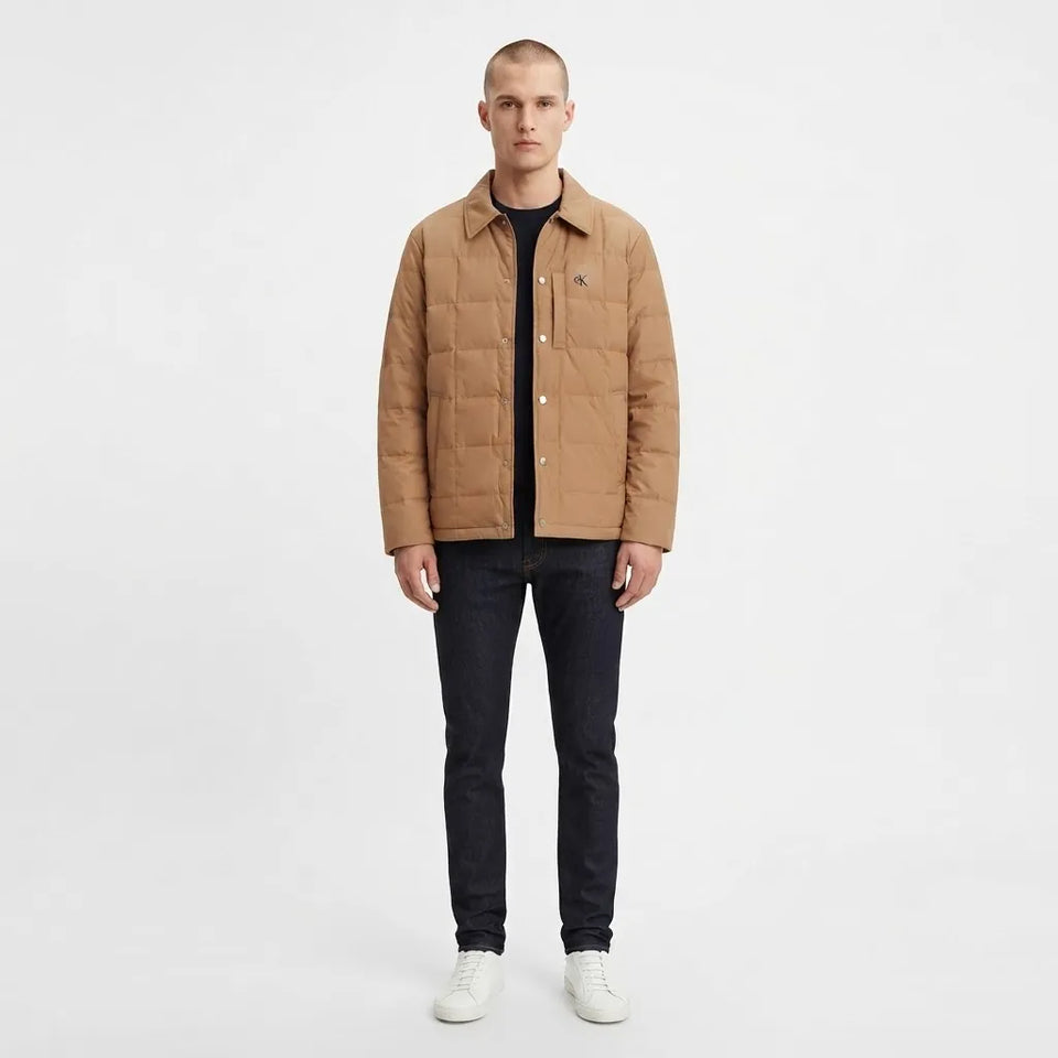 A man modeling a tan quilted jacket over a black shirt and dark jeans, standing against a plain white studio background.