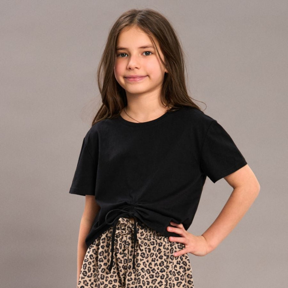 A young girl modeling a black short-sleeve t-shirt and leopard print shorts against a solid gray studio background.