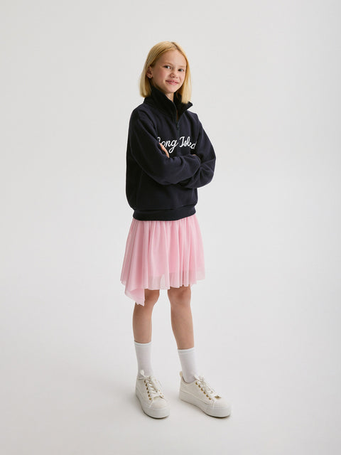 Young girl wearing a black hoodie with text, pink skirt, white socks, and white sneakers on a light gray background