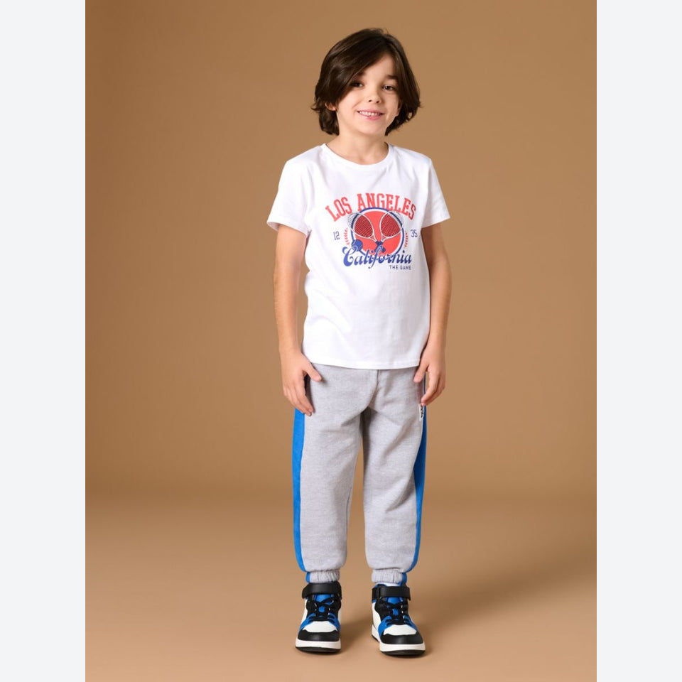 A young boy modeling a white graphic t-shirt and light gray sweatpants with blue side stripes, standing against a solid tan studio background.