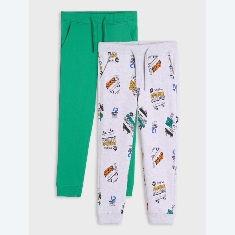 Two pairs of children's jogger sweatpants laid flat on a light gray background. The back pair is solid bright green, and the front pair is light heather gray with an allover colorful graphic print featuring cartoon vehicles and text.
