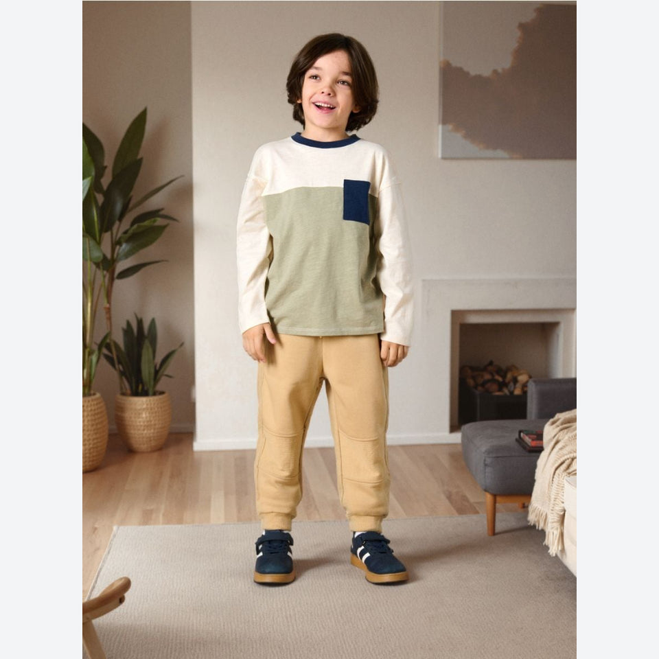 A young boy stands in a cozy living room wearing a color-blocked long-sleeve shirt with cream, sage green, and navy panels, paired with tan pants and navy sneakers.