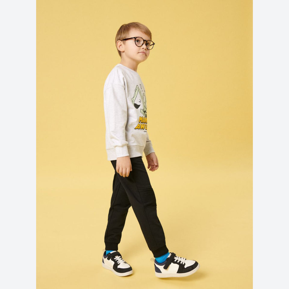 A young boy wearing round black glasses, a light gray graphic sweatshirt, black pants, and black-and-white sneakers with bright blue socks, standing against a solid yellow studio background.