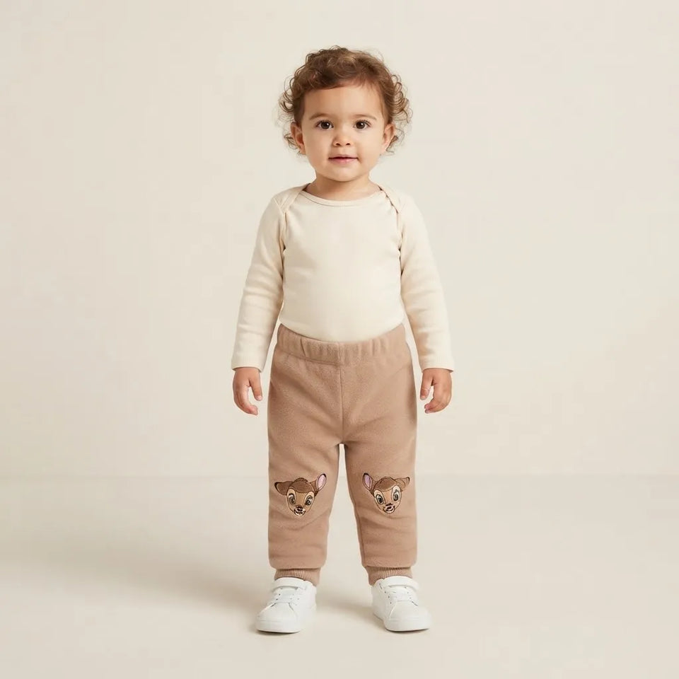 A toddler standing against a warm beige studio background, wearing a cream long-sleeve top and tan jogger pants with embroidered animal faces near the lower legs, paired with white sneakers.
