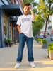 A young boy stands on a city sidewalk wearing a white graphic t-shirt with 'WEEKEN' printed in blue, blue jeans, and white sneakers.