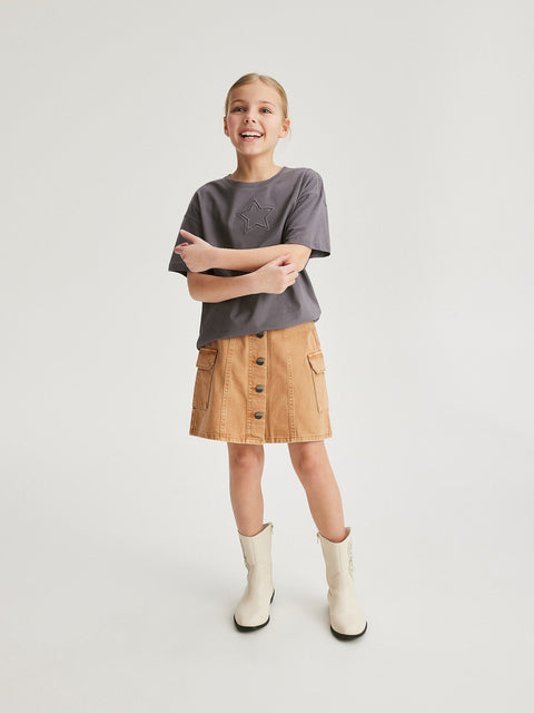 Young girl wearing a gray t-shirt and tan skirt with buttons on a white background