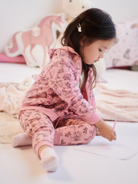 Child in pink pajamas sitting on a bed with pink pillows and blanket.