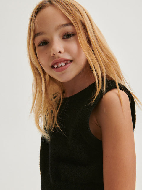 Young girl with blonde hair wearing a black sleeveless top against a plain background