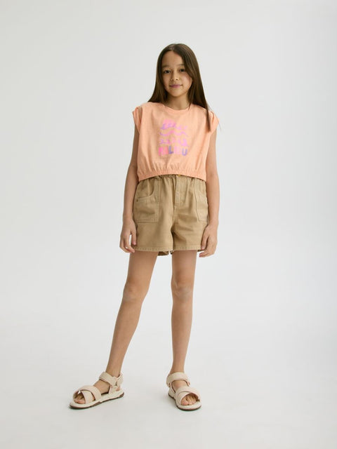 Young girl wearing a sleeveless pink top and beige shorts on a white background