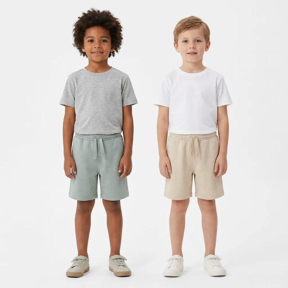 Two children standing side by side wearing matching short-sleeve t-shirts and drawstring shorts. The child on the left wears a heather gray t-shirt with sage green shorts, and the child on the right wears a white t-shirt with beige shorts.