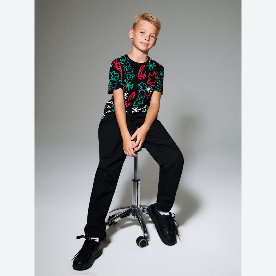 A young boy sits on a metal stool in a studio setting, wearing a short-sleeve crewneck t-shirt with a vibrant green, red, and white paisley pattern, paired with black pants and black sneakers.