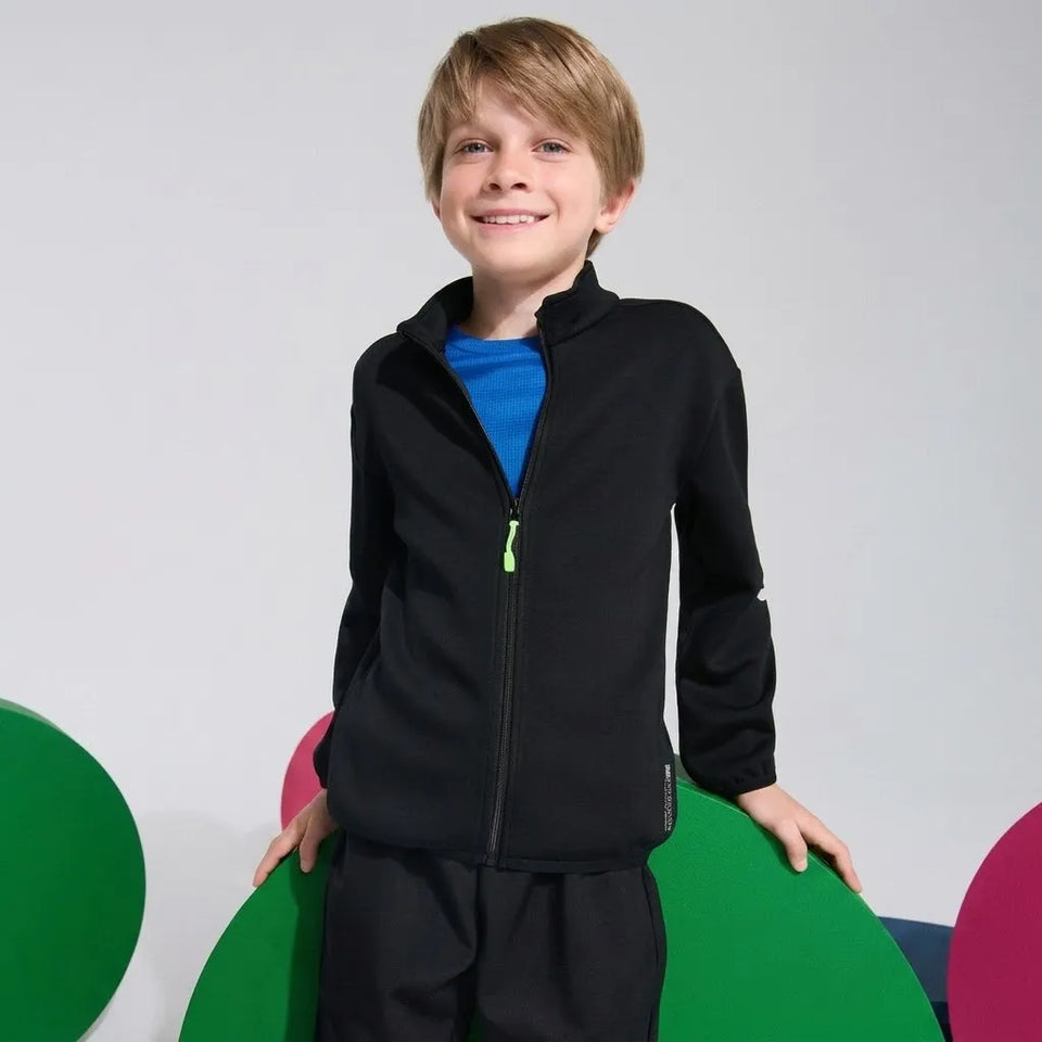 A young boy smiling and wearing a black zip-up fleece jacket over a bright blue t-shirt, standing in front of a light gray background with colorful green and pink circular props.