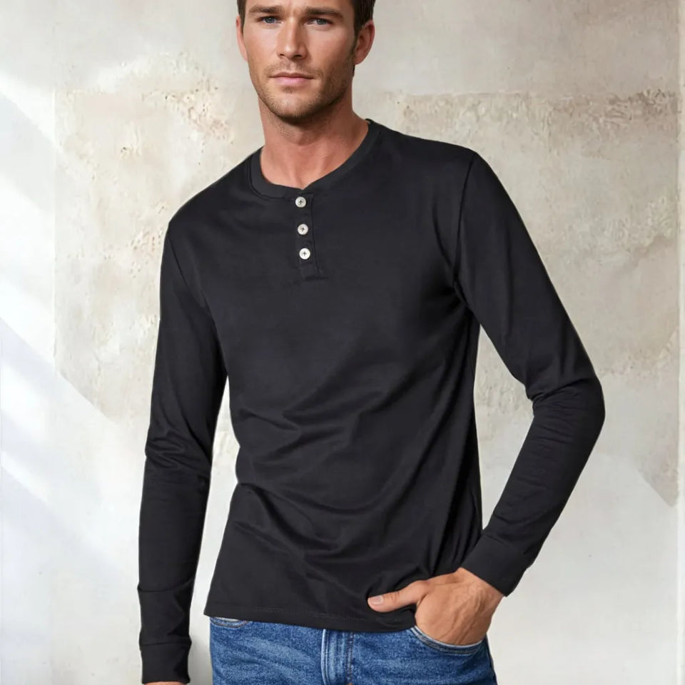 A man wearing a black long-sleeve henley shirt with three buttons on the collar, paired with blue jeans, standing against a textured beige wall.