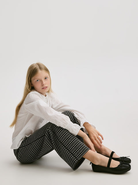 Young girl sitting on a white floor wearing a white shirt, checkered pants, and black shoes.