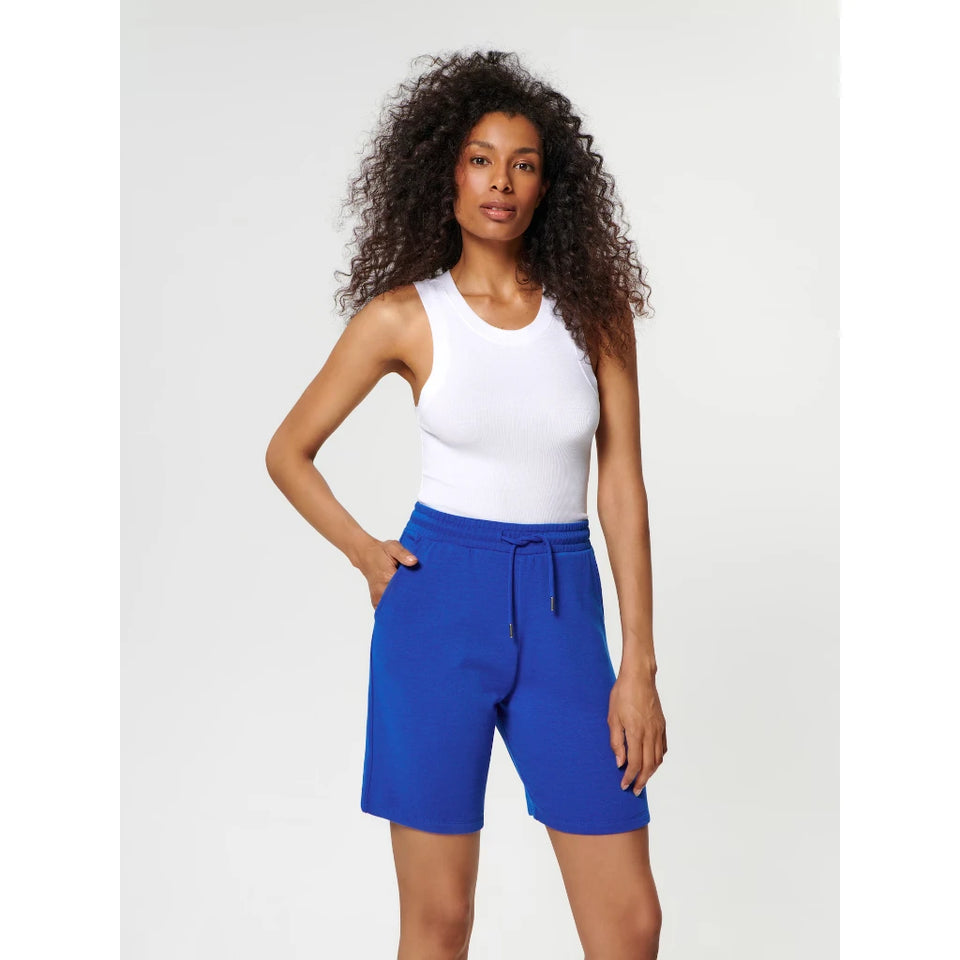 A woman modeling bright royal blue athletic shorts with a drawstring waist, paired with a white tank top, against a light gray studio background.