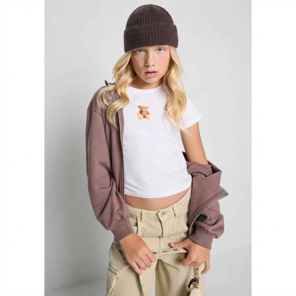 A young girl modeling a white crop top with a teddy bear graphic, styled with a brown knit beanie, a brown zip-up jacket, and beige cargo pants against a light gray studio background.