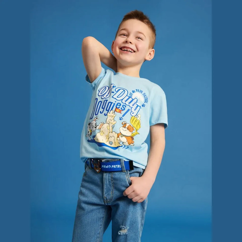 A young boy smiling and posing in a light blue graphic t-shirt and blue jeans against a solid blue studio background.