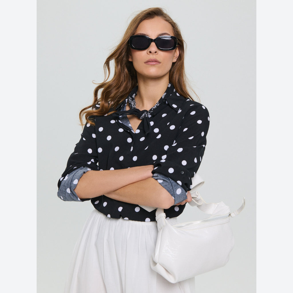 A woman wearing a black long-sleeve blouse with white polka dots and a keyhole cutout, styled with a white skirt, black sunglasses, and a white shoulder bag against a light gray studio background.