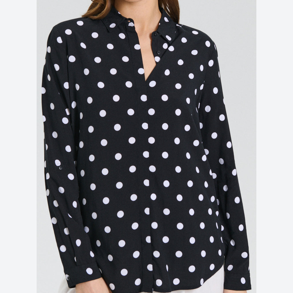 A woman wearing a black long-sleeve button-up blouse with a white polka dot pattern, shown from the neck down against a light gray studio background.