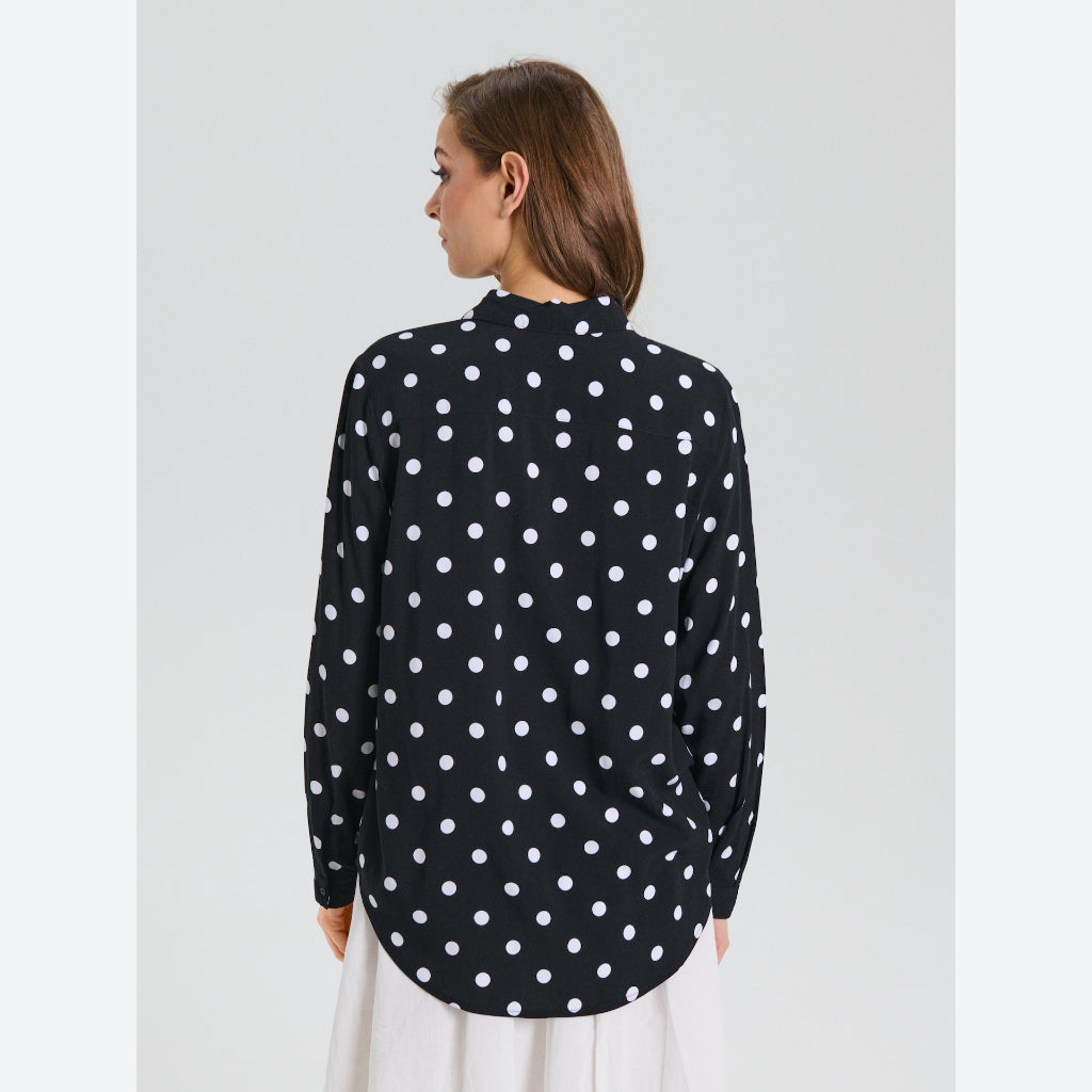 A woman viewed from the back wearing a black long-sleeve blouse with a white polka dot pattern and a white skirt, set against a plain light gray studio background.