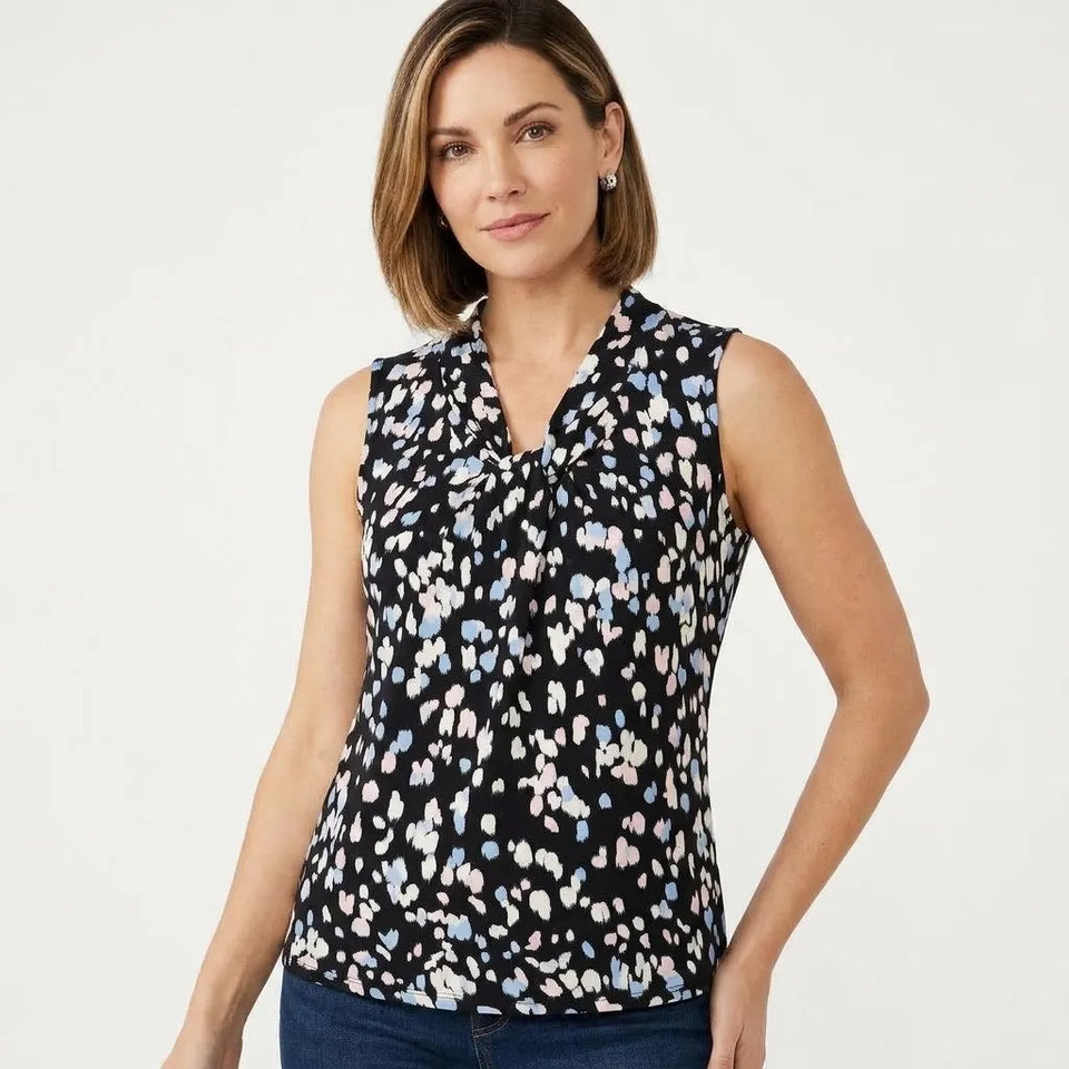 A woman modeling a sleeveless black blouse with a white, light blue, and pink abstract pattern, paired with dark blue jeans, against a plain white studio background.