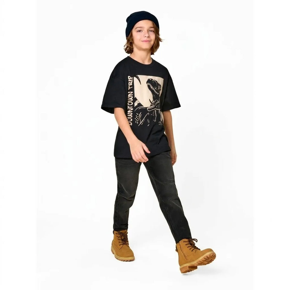 A child model wearing a black graphic t-shirt with a beige and black print, dark gray pants, tan boots, and a black beanie, standing against a plain white background.