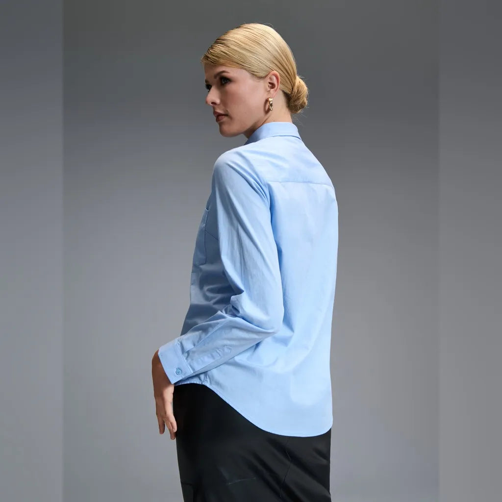 A woman wearing a light blue long-sleeve button-up shirt and black trousers, photographed from the back against a neutral gray studio background.