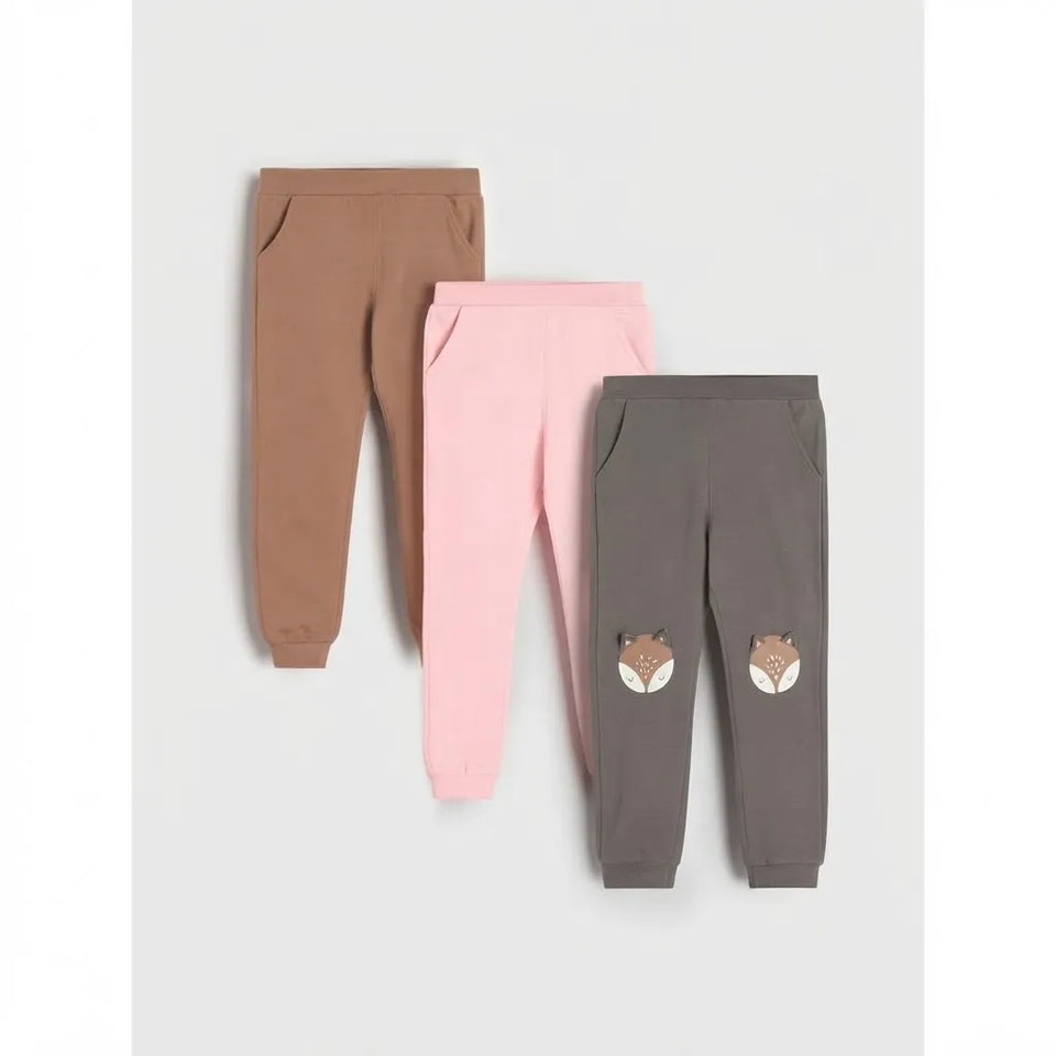 Three pairs of children's jogger pants in brown, pink, and dark gray, arranged overlapping on a light gray background. The dark gray pair features two embroidered fox faces on the lower legs.