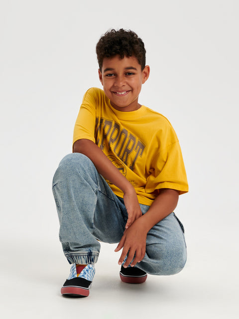 Child wearing a yellow t-shirt with text and blue jeans on a white background