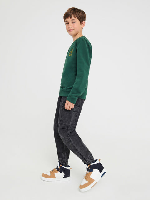 Child wearing a green sweatshirt with a logo and dark jeans on a white background