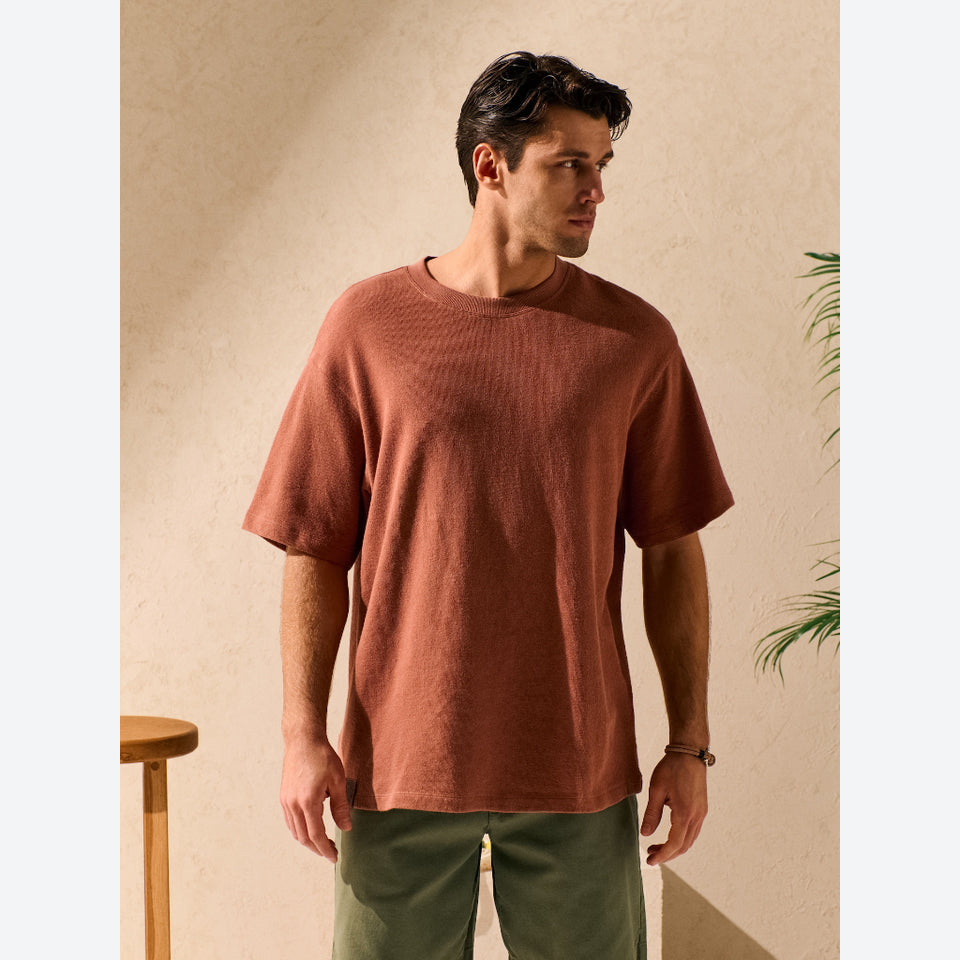 A man wearing a textured, short-sleeve crew-neck t-shirt in a warm rust or terracotta color, paired with olive green pants, standing against a textured beige wall.