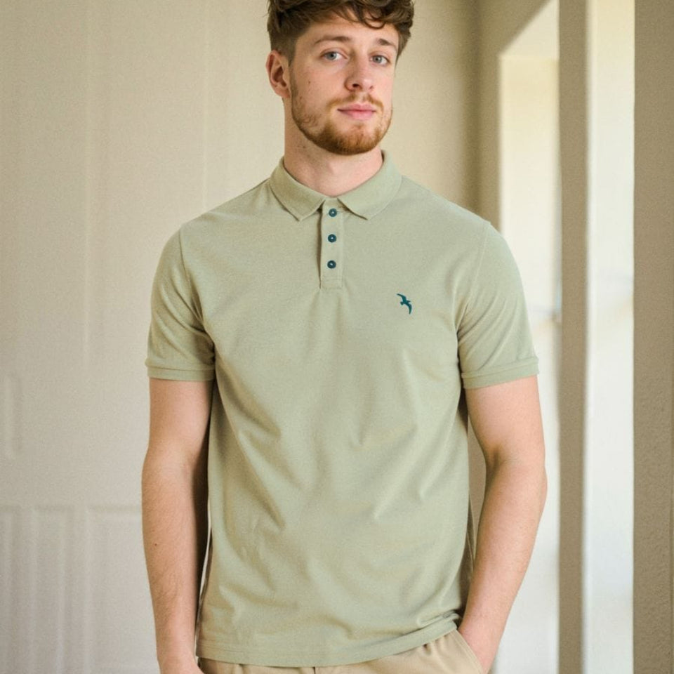 A man wearing a light sage green polo shirt with a small embroidered bird logo on the chest, paired with khaki cargo pants, standing in a brightly lit indoor setting.